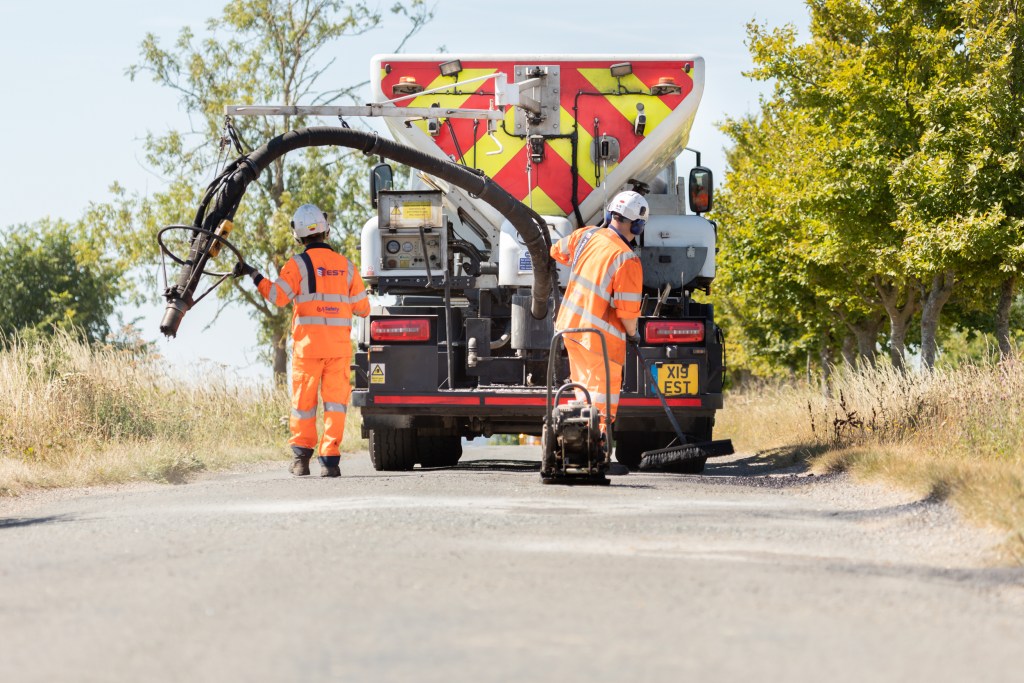 Two men spray injection patching on a country road in Milton Keynes. One man is holding a long black tube attached to the tanker and one man is brushing the tarmac just been put on the road